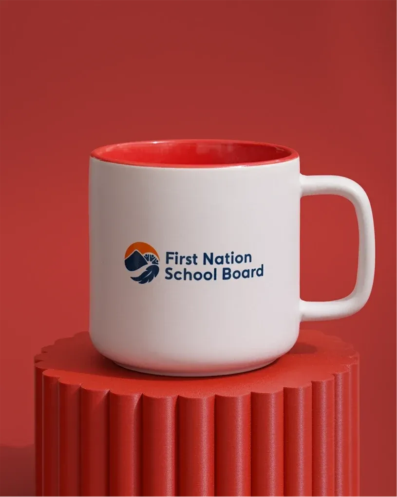 White ceramic mug with a red interior and the First Nation School Board logo on the front.